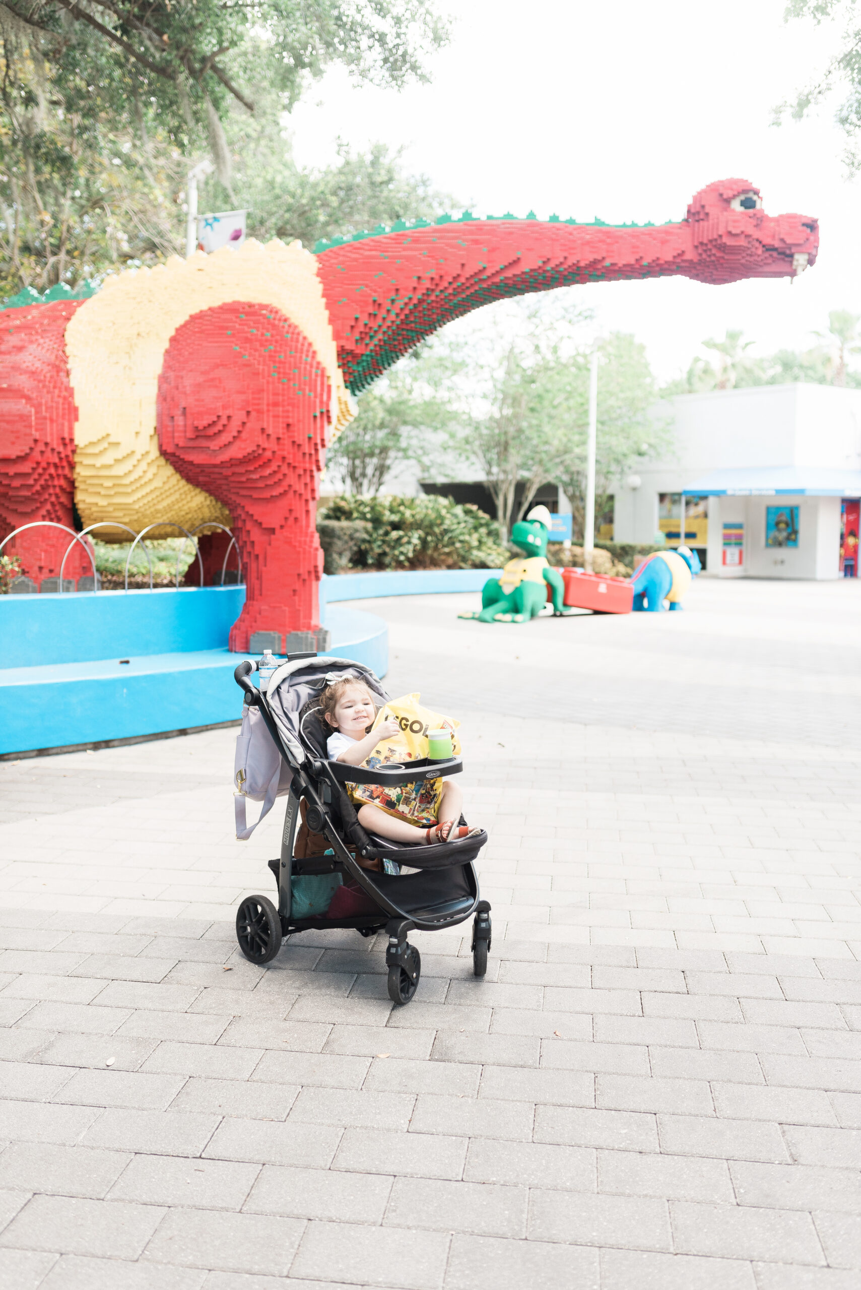 Little girl in stroller holding LEGOLAND bag sitting below a giant LEGO dinosaur
