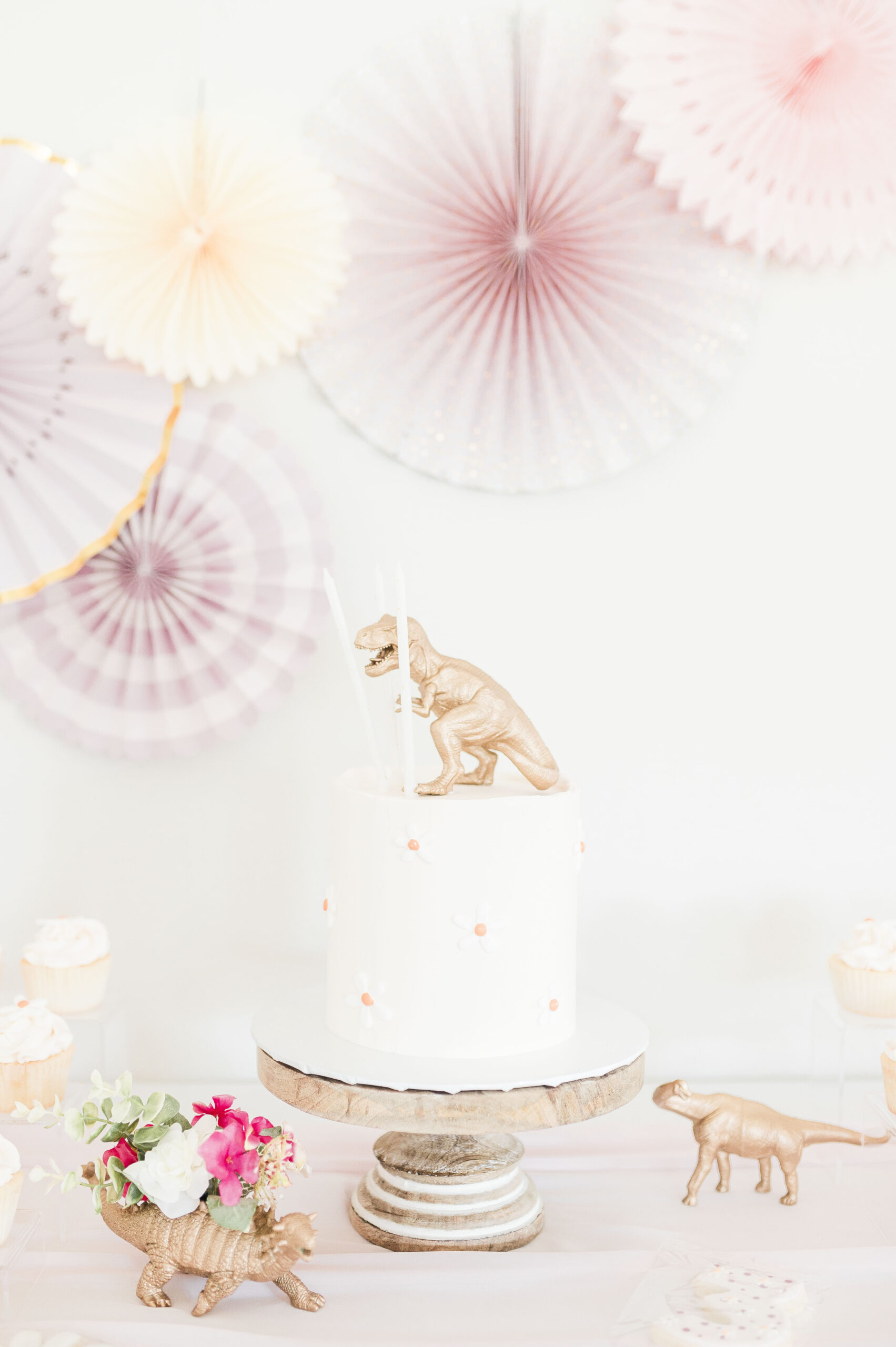 Cake for three rex birthday for girl, round cake with frosted daisies around it with gold t-rex on top with candles, flowers and cupcakes surround the cake. dinosaur-themed birthday