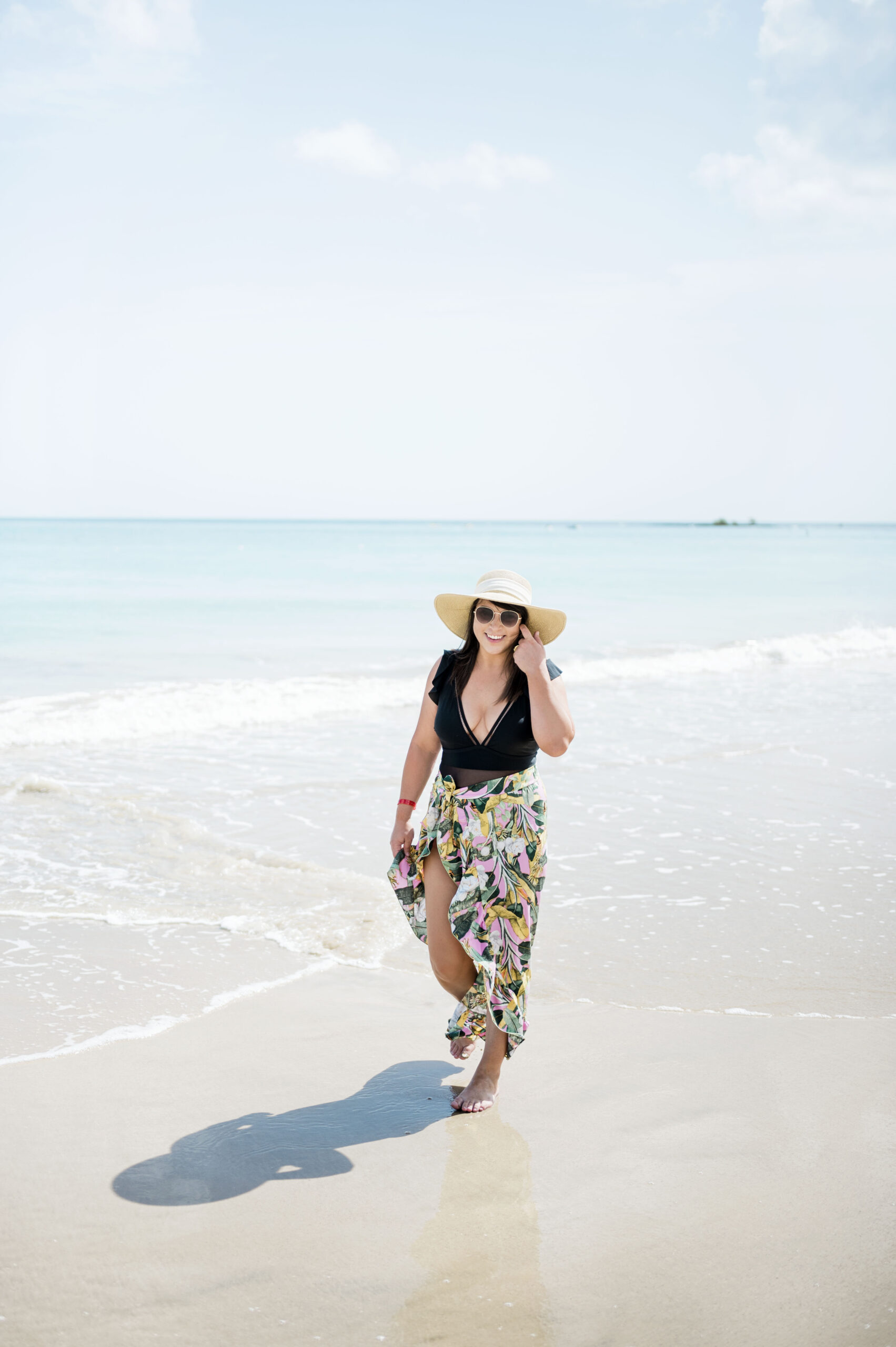 female in black one piece swim suit and pink tropical sarong walking on the beach 