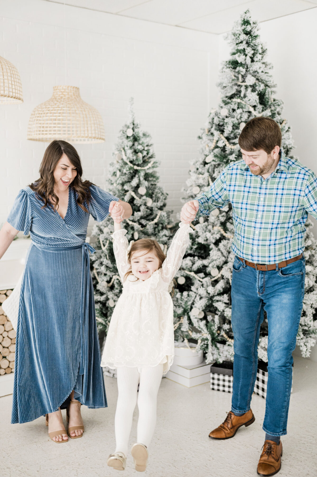 Brittney Naylor in maxi blue velvet dress, holding daughters hand, and husband John holding other hand of daughter wearing jeans and shirt, in front of fireplace and christmas tree