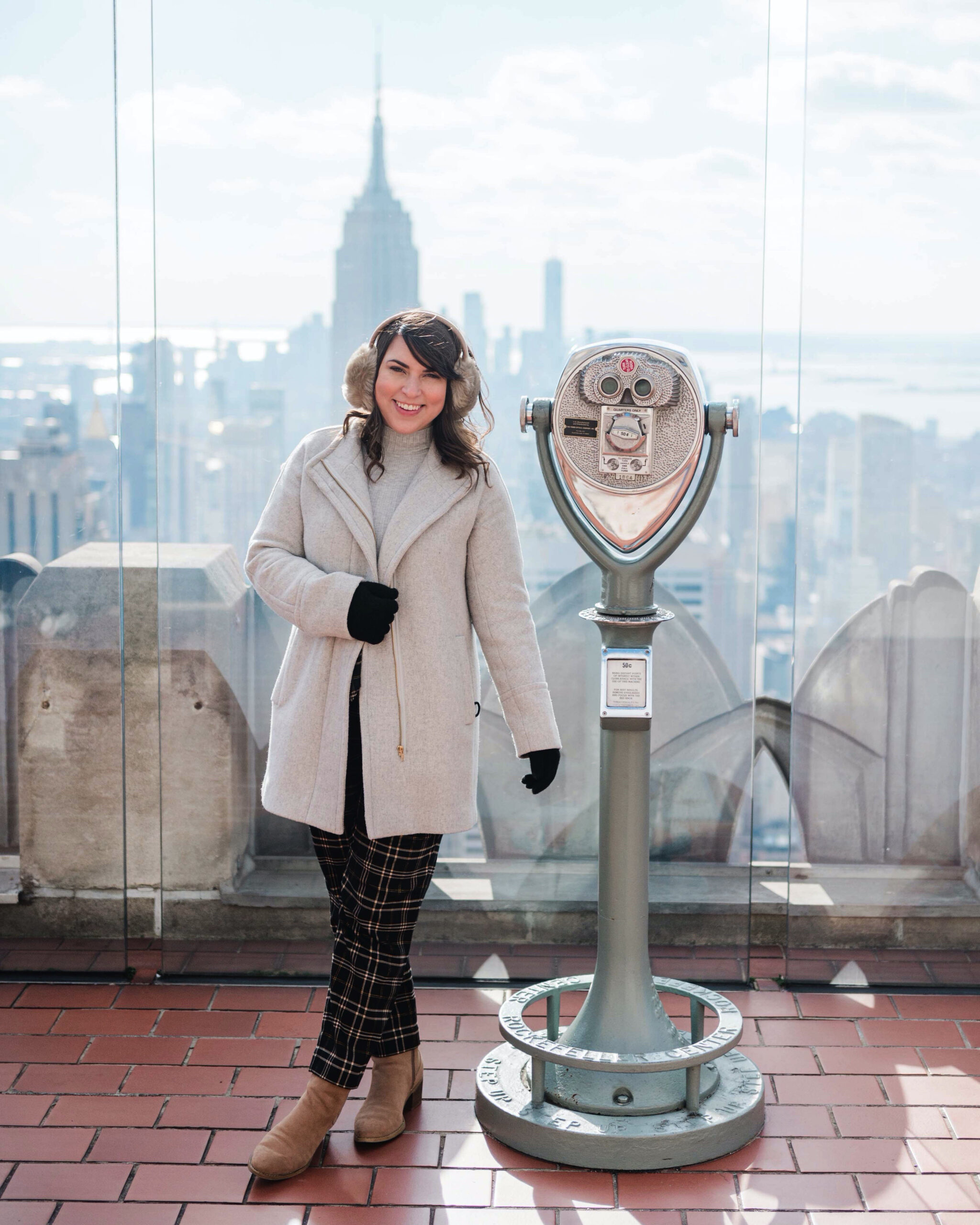 Brittney Naylor wearing black plaid pants, brown boots, grey petticoat, black gloves, and ear muffs on top of the rockefeller plaza with views of the empire state building and other sky scrapers in New York City. NYC Christmastime Bucket List Things to do.