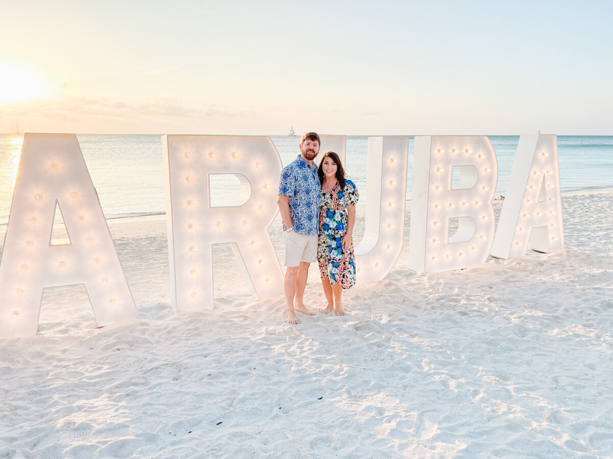 Couple standing on the beach in front of large marquee letters spelling Aruba