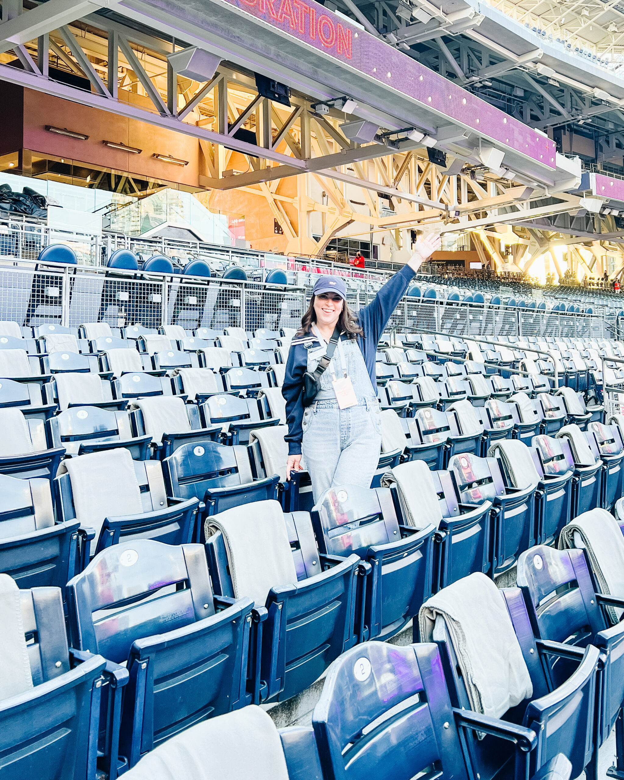 Brittney Naylor wearing overalls and ball cap with hands in the air standing in the stands at Petco Park in San Diego