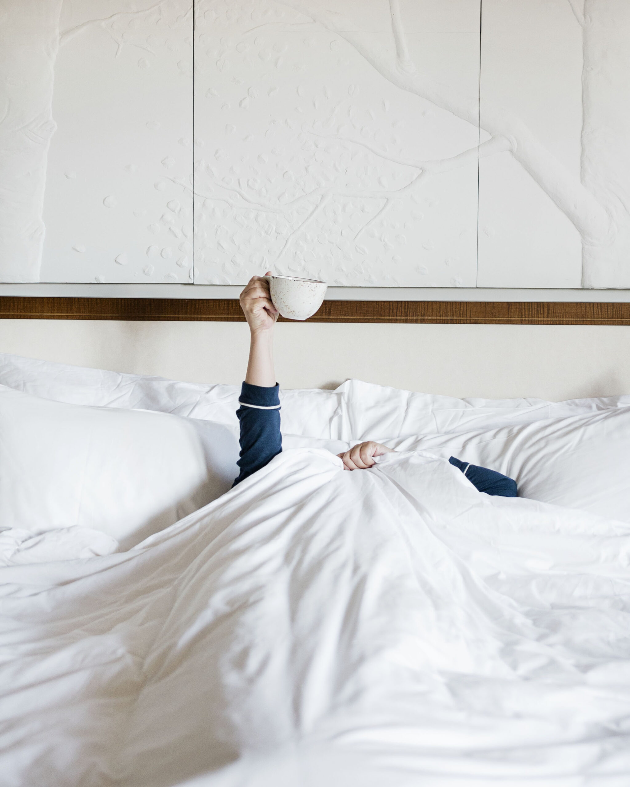 brittney naylor laying under the covers in bed at the four seasons jackson hole, holding a coffee mug up in the air