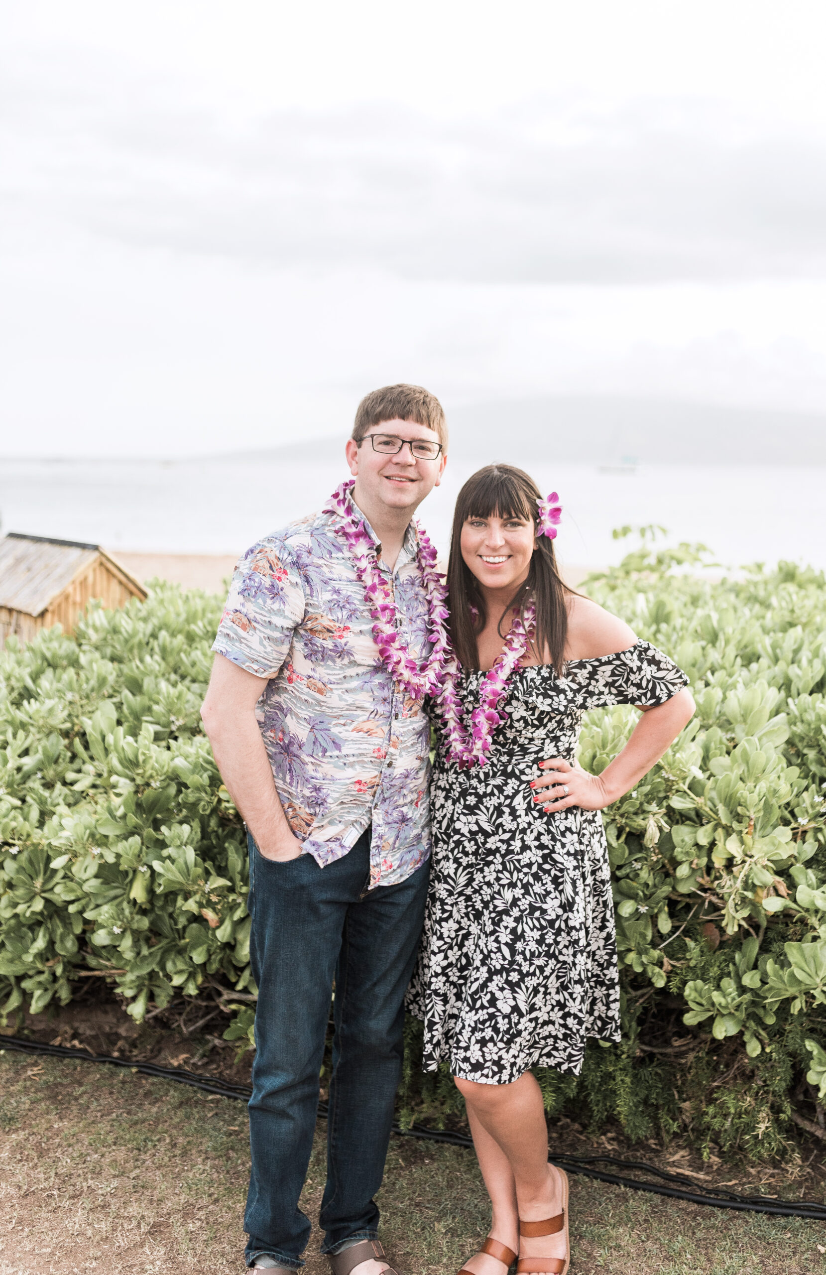 John and Brittney Naylor posed together in Maui Hawaii at Luau. Both are wearing purple flower leis and are standing in front of greenery with ocean behind. Maui with adventure.