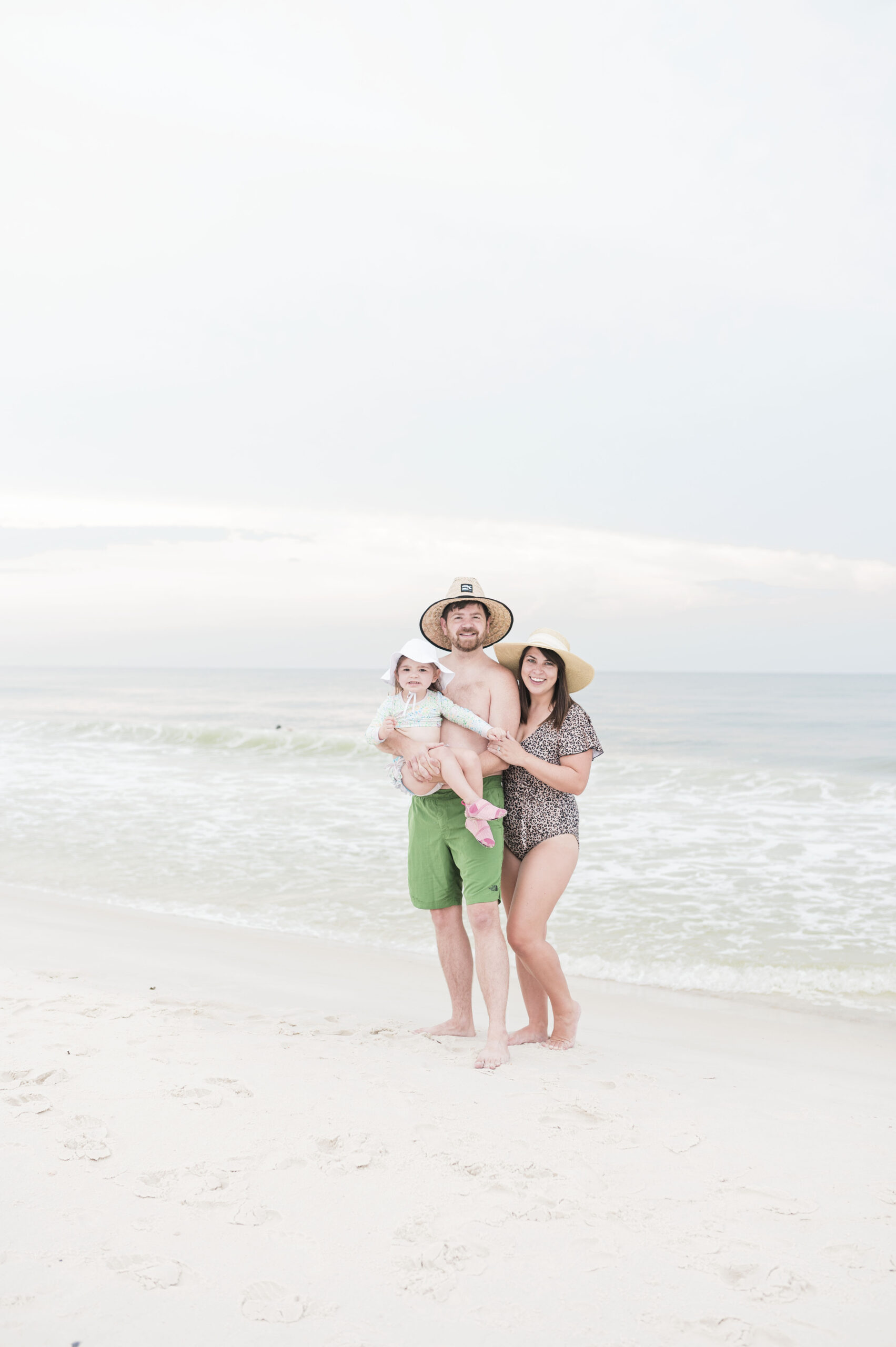John, Brittney, and Eleanor Naylor standing together in swim suits on the beach in gulf shores, alabama. Traveling on a budget.