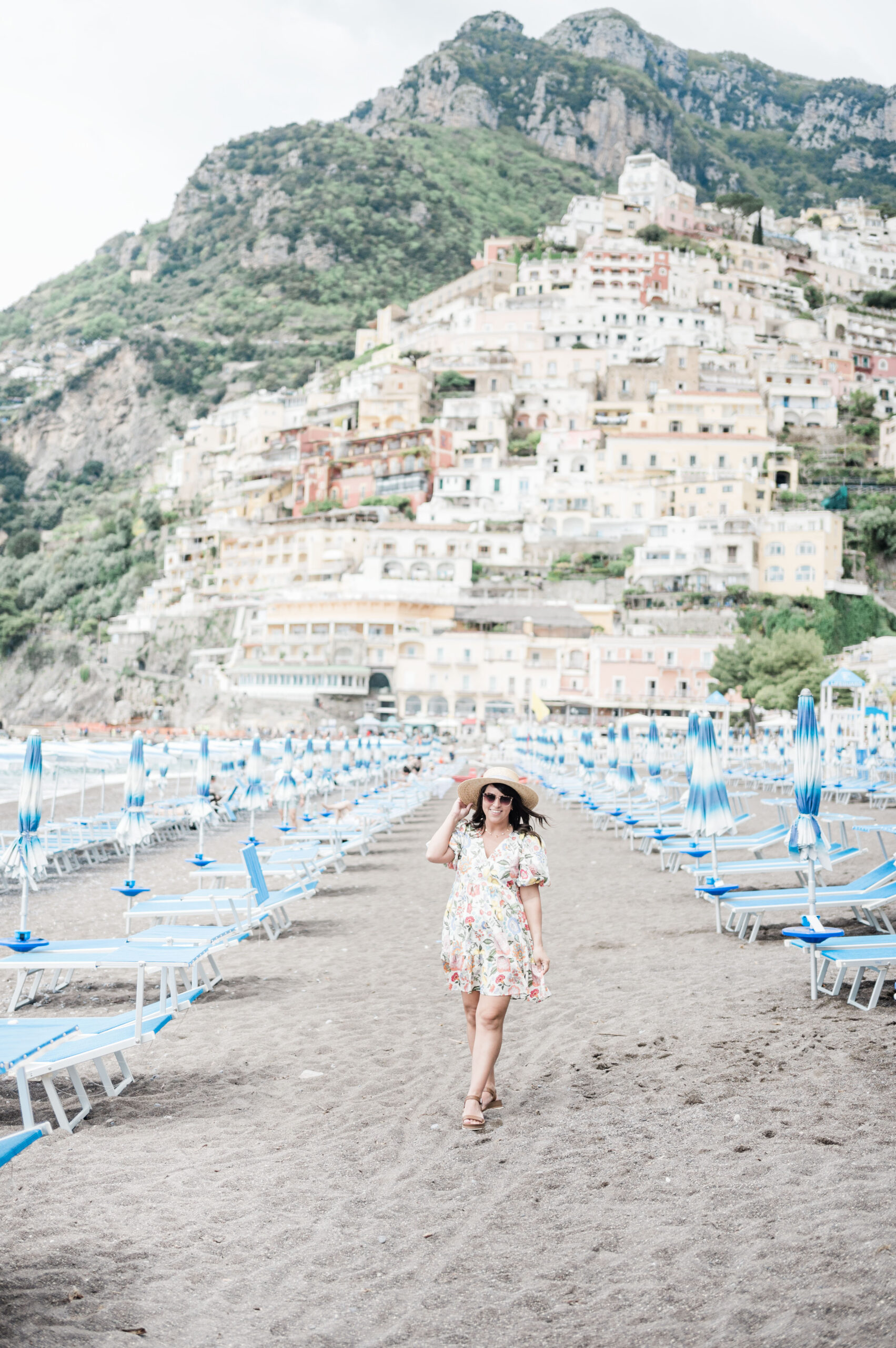 Brittney Naylor standing between blue beach chairs on the beach in Positano Italy. A 10 day Italy Itinerary as First Timers. A Mom Well Traveled Blog.