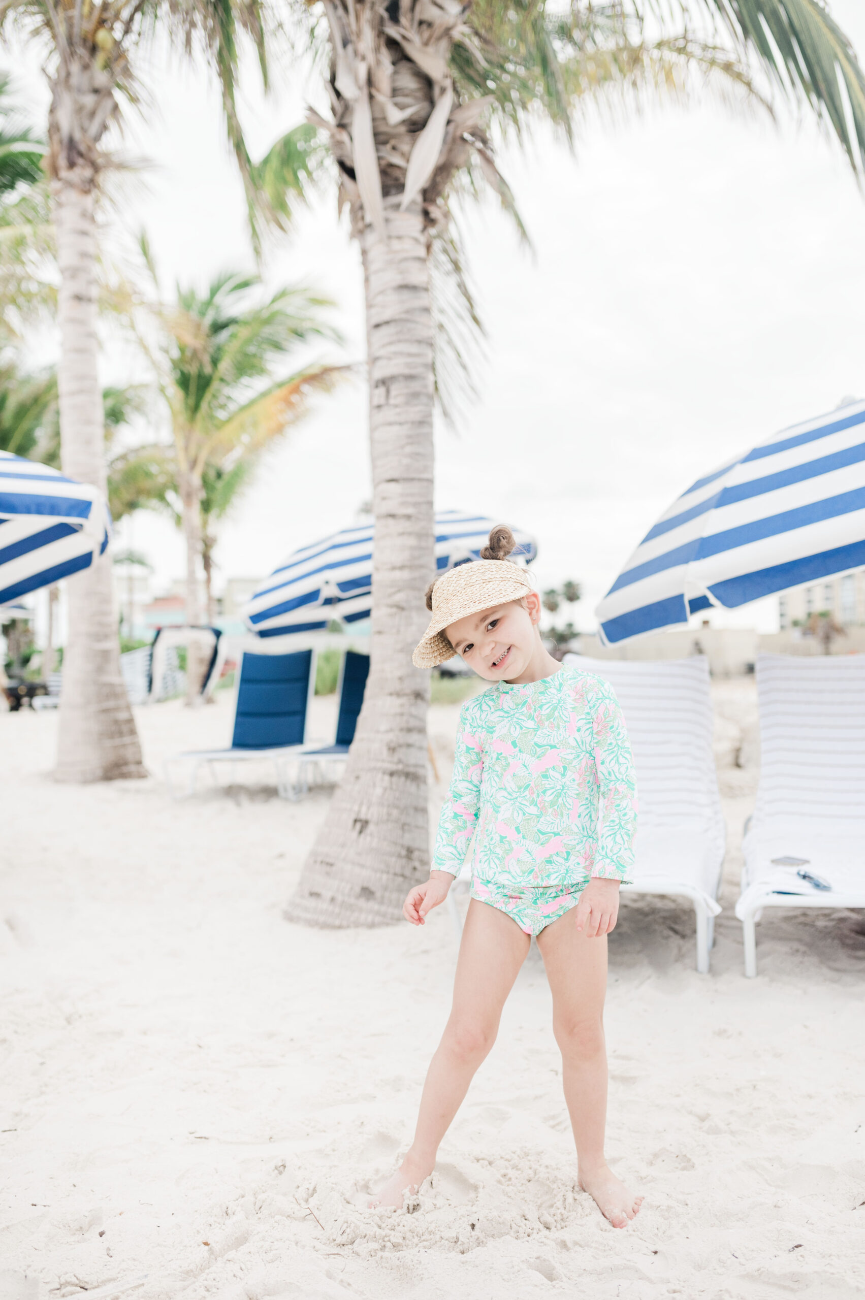 Eleanor in Lilly Pulitzer rashguard swimsuit standing on the beach at JW Marriott Clearwater Beach with white and blue striped umbrellas behind her