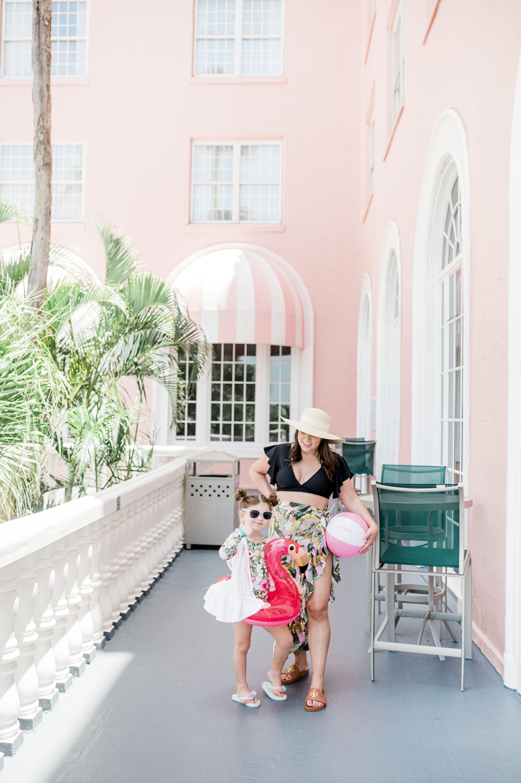 Brittney Naylor looking at daughter. Both are in swimwear. Brittney is holding a beach ball while her daughter had a pink flamingo float around her waist. Her daughter is holding up a thumbs up as Brittney looks on. Both are standing on Balcony at the Pink Palace Instagrammable hotel in Florida