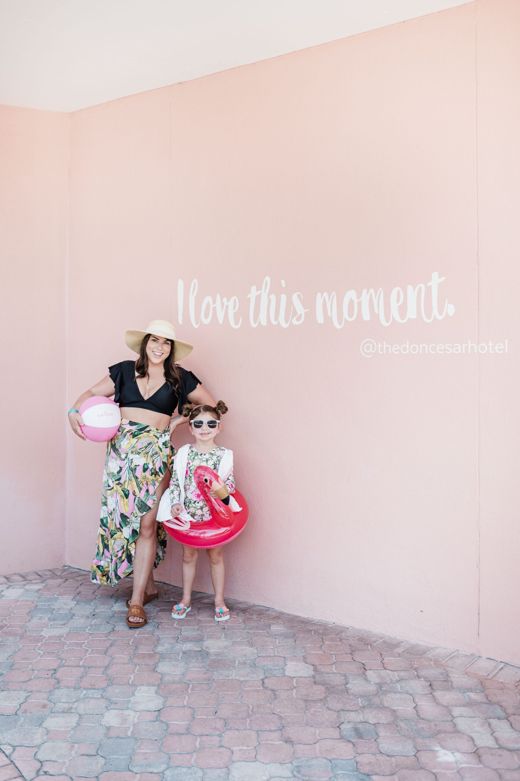 Brittney Naylor and daughter in swimwear, flamingo float, and beach ball standing in front of mural that says I love this moment at The Don CeSar in Saint Pete Pink Palace