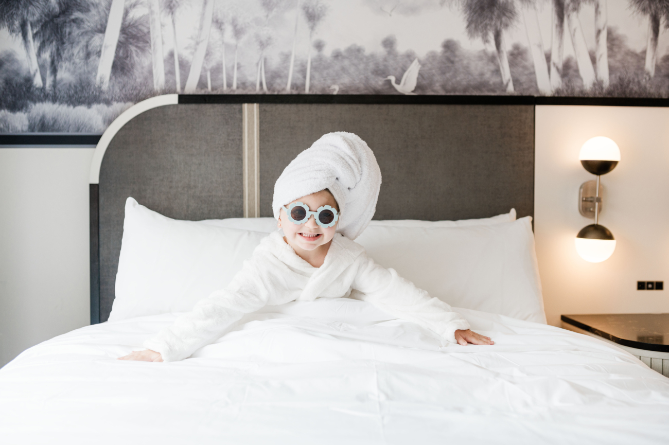 Eleanor in bed at the Caribe Royale in Orlando. She is wearing a white robe and white towel on her head, as well as, blue sunglasses. Orlando Staycations.