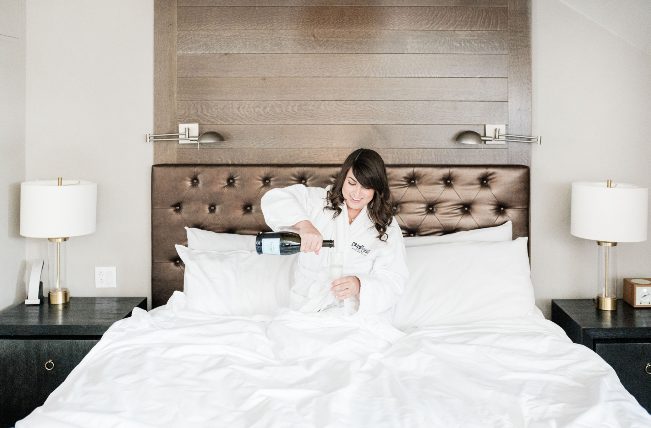 Brittney Naylor pouring white whine in a glass while wearing a robe from The Crawford Hotel and sitting in bed. Denver Hotels.