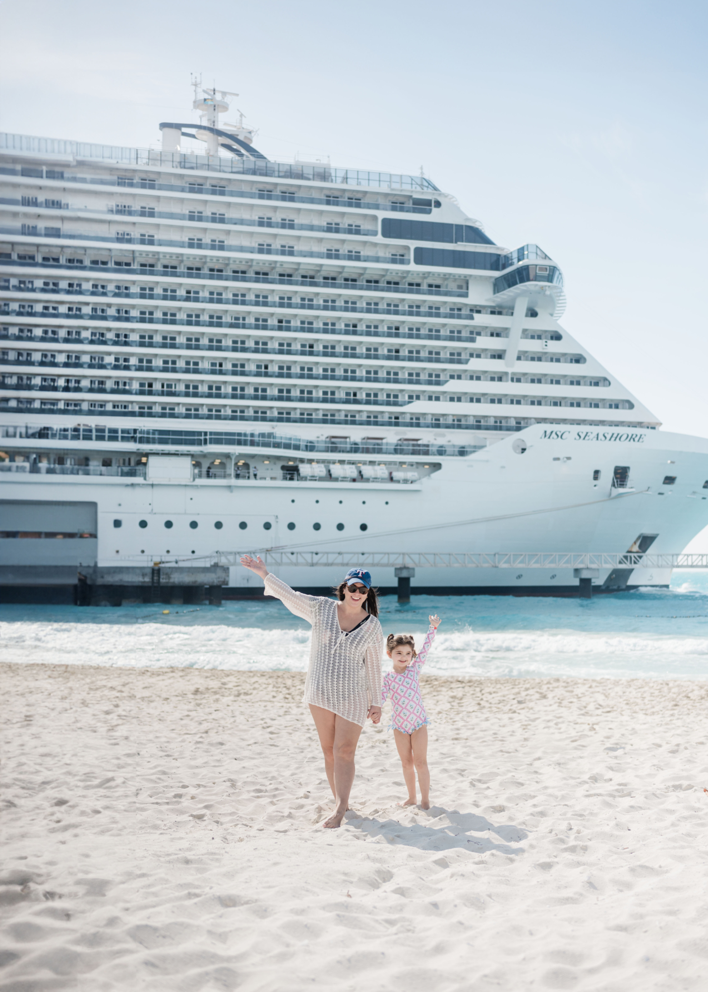 Eleanor and Brittney raising their hands as they stand in the sand at Ocean Cay in front of the MSC Seashore. MSC vs Royal Caribbean: what cruise is best for families.