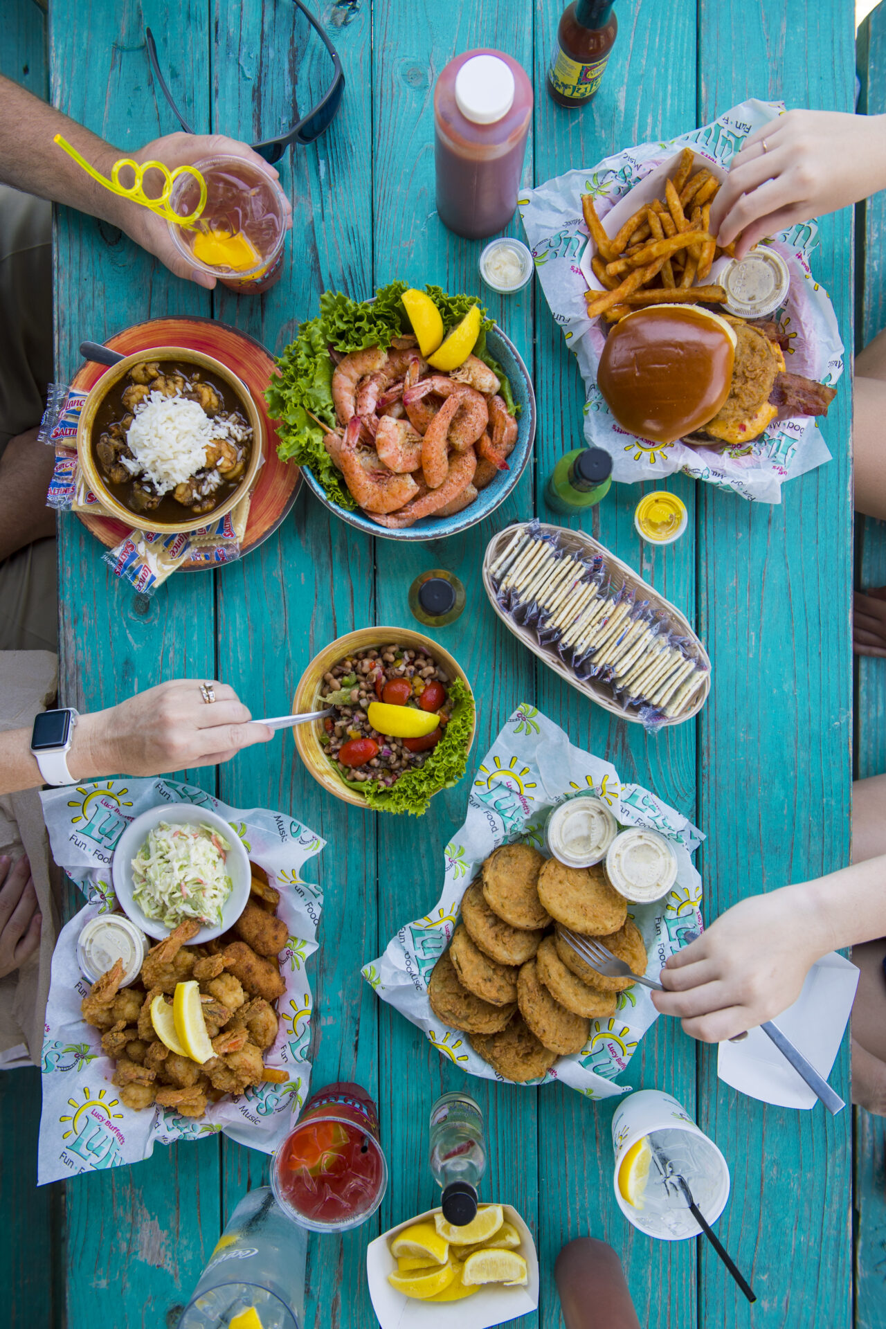 overhead photo of a variety of foods on teal table at LuLu's restaurant in Gulf Shores, Alabama. Food includes fried green tomatoes, fried shrimp, a sandwich, and more. Photo provided by Visit Alabama Beaches. Photo courtesy of Gulf Shores & Orange Beach Tourism