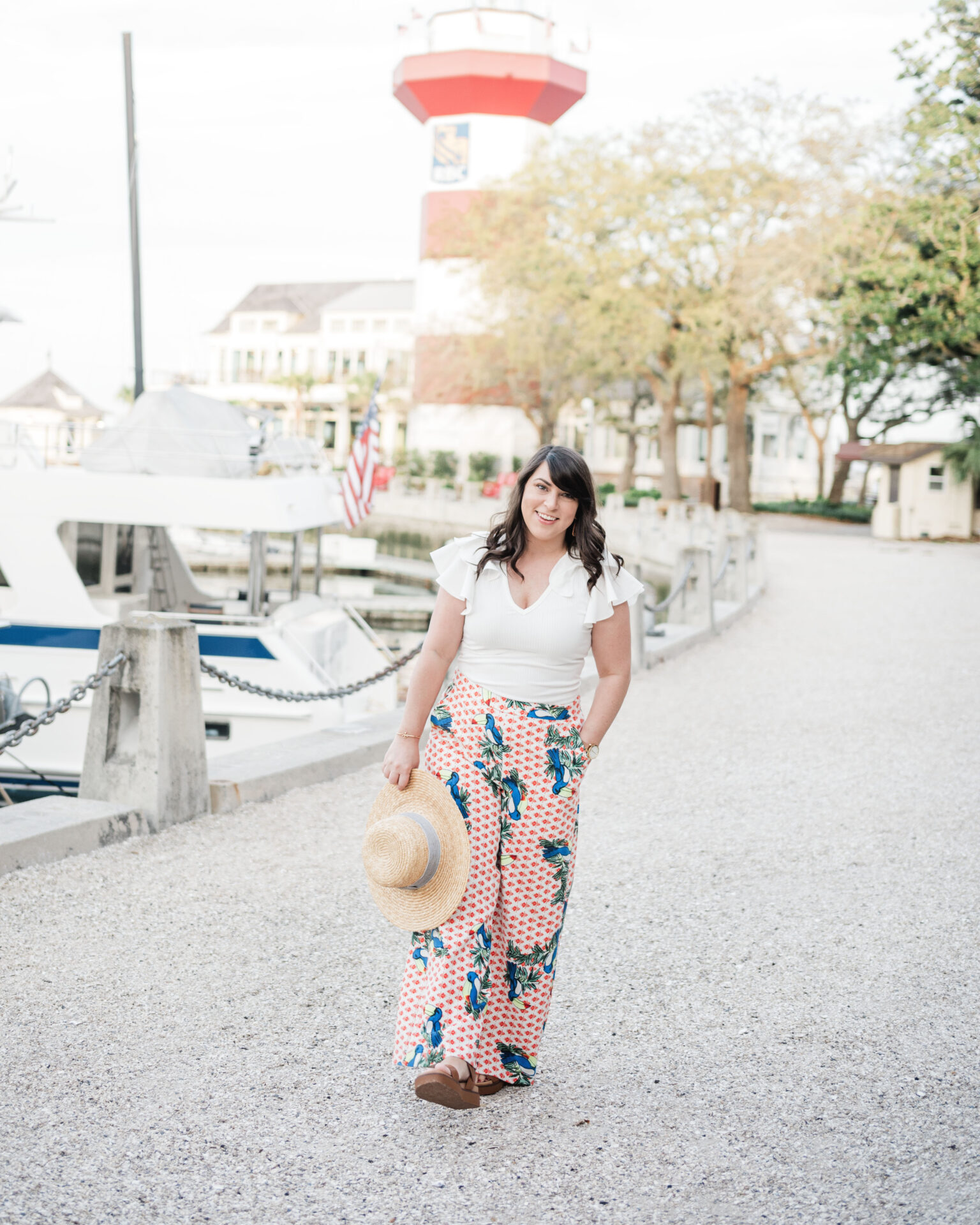 Brittney Naylor wearing tropical pants and white blouse walking through Harbour Town in Hilton Head Island. Harbour Town Lighthouse is in the background and boats are to the side in the water.
