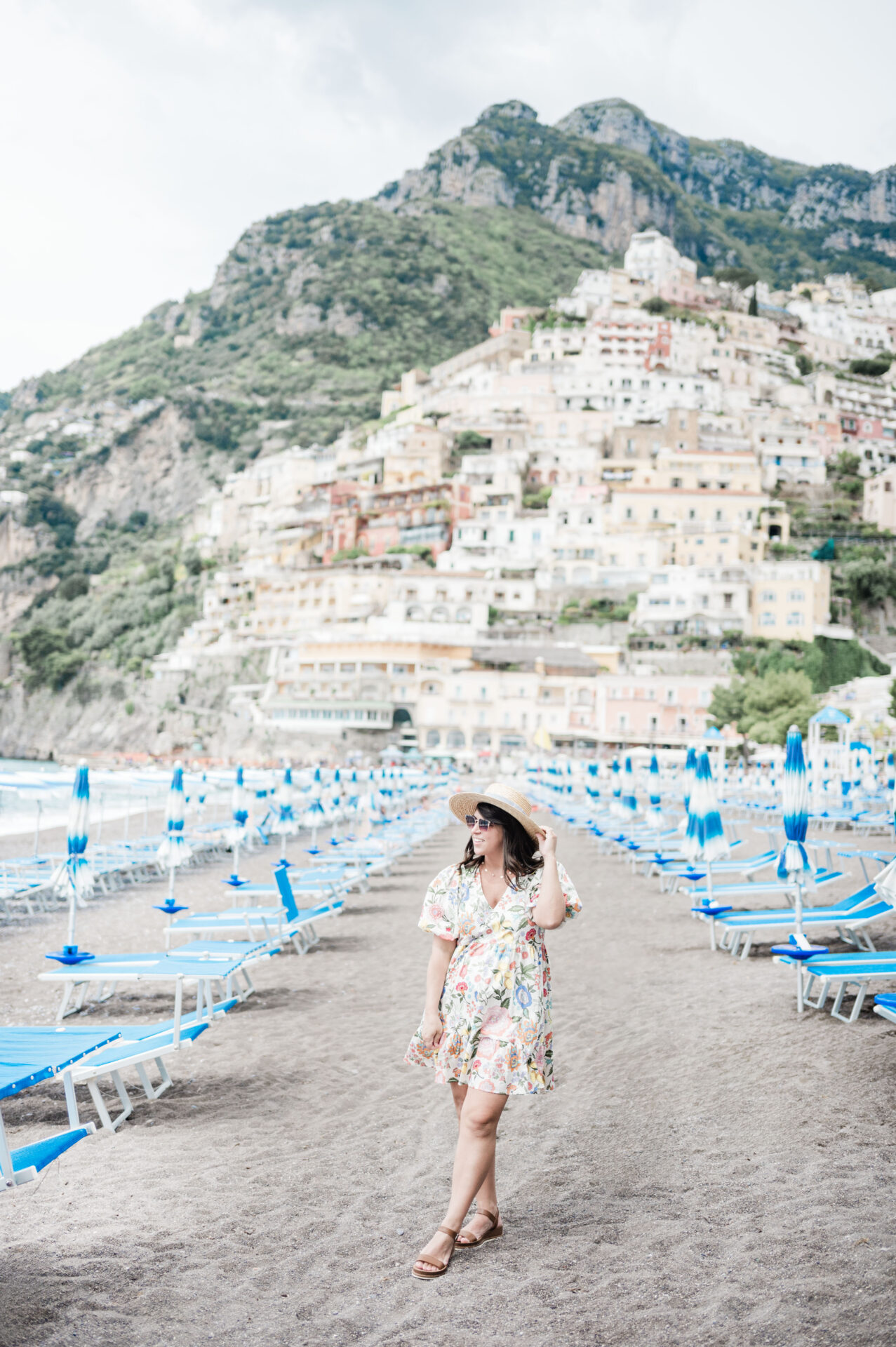 Brittney Naylor walking between beach chairs on the beach in Positano wearing a dress, looking to the side and holding onto hat. Positano hillside with buildings in the background. How to spend 3 days in Positano.