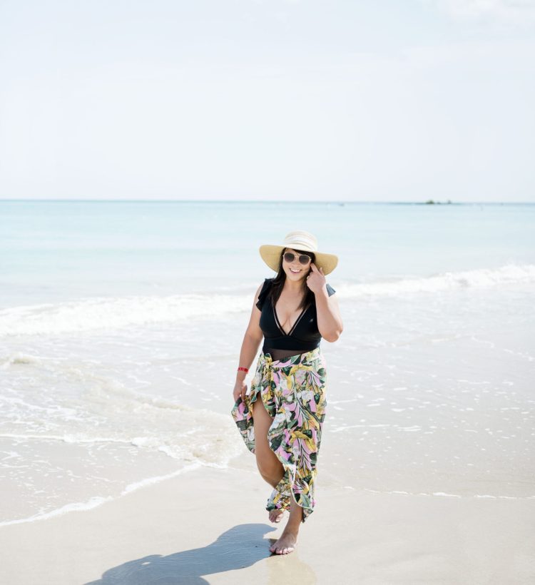 Brittney Naylor of A Mom Well Traveled in black one piece swim suit and pink tropical sarong walking on the beach, helping you find your next vacation destination